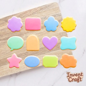 May include: A collection of twelve sugar cookies with smooth, colorful icing in various shapes including a star, heart, speech bubble, and geometric forms. The cookies are arranged in rows, featuring shades of purple, pink, blue, yellow, green, and orange.