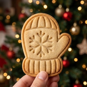 May include: A light brown, mitten-shaped cookie featuring an embossed snowflake design in the center and ribbed texture patterns on the cuff and top sections. The cookie has a smooth, baked surface with defined edges and a consistent golden-brown color.