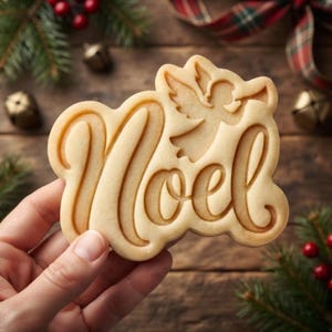 May include: A light beige cookie cutter shaped like the word Noel in a cursive script font, featuring an embossed angel figure integrated into the letter O. The cutter has a smooth, molded surface with raised edges for creating detailed impressions.