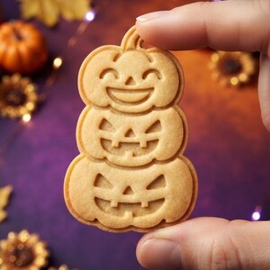 May include: A light tan, baked cookie shaped like a vertical stack of three pumpkins. The top pumpkin features a smiling face with curved eyes, while the middle and bottom pumpkins display carved jack-o'-lantern faces with triangular eyes and mouths.