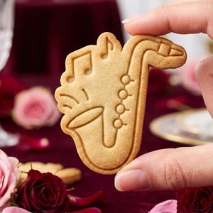 May include: A light brown, baked cookie shaped like a saxophone with embossed musical notes and circular keys. The cookie has a smooth, golden-brown surface and a distinct outline that follows the shape of the instrument, showing clear details.