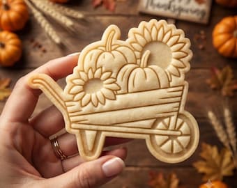 May include: A light beige sugar cookie shaped like a wheelbarrow filled with two pumpkins and two sunflowers. The cookie features embossed details showing the wooden slats of the wheelbarrow, the spokes of the wheel, and the textures of the flowers.