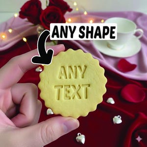 May include: A round, yellow cookie with a scalloped edge, imprinted with the words "ANY TEXT". The cookie is held in a hand, with a banner above it that reads "ANY SHAPE". The background includes roses and a teacup.