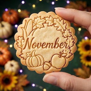 Peut inclure: Un biscuit rond à bords festonnés présentant un motif en relief avec le mot November entouré d'une couronne de feuilles d'automne, d'une citrouille, d'un gland et d'une pomme. Le biscuit a une couleur brun doré clair et une texture lisse.