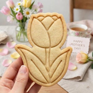 May include: A tulip-shaped cookie with detailed lines, held in a hand. The cookie is light brown and sits in front of a vase of flowers, a teacup, and a card that reads "Happy Mother's Day!"