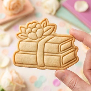 May include: A light beige cookie shaped like a stack of two books topped with a large flower. The cookie features embossed lines that define the pages, covers, and petals of the floral design, creating a clear and detailed raised texture.