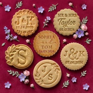 May include: A collection of personalized cookies with dates and initials, arranged on a burgundy surface with floral accents. The cookies feature embossed text, including names and wedding dates, and are surrounded by small flowers and greenery.