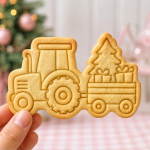 May include: A light tan, baked sugar cookie shaped like a tractor pulling a trailer. The trailer carries a Christmas tree and two gift boxes. The cookie features embossed outlines of the tractor, wheels, tree, and gifts on its smooth surface.