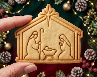 May include: A light brown, house-shaped cookie featuring an embossed nativity scene. The design depicts two figures kneeling on either side of a central manger with a baby, all beneath a large star at the peak of the roof-shaped cookie structure.
