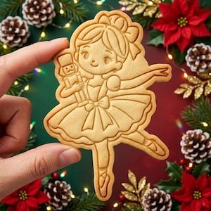 May include: A light tan cookie shaped like a ballerina holding a nutcracker. The cookie features embossed lines depicting a dancer in a tutu with a bow in their hair, holding a small nutcracker figure, with defined ballet slippers.