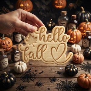 May include: A light-colored, flat cookie cutter shaped like the words 'hello fall' in a cursive script, featuring an embossed maple leaf icon and a heart outline. The design is made of a smooth, light-toned material with raised edges for stamping.