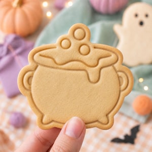 May include: A light tan, cauldron-shaped sugar cookie with embossed details showing a bubbling liquid spilling over the rim. The cookie features two side handles, a rounded base with small feet, and three circular bubbles on the top surface.