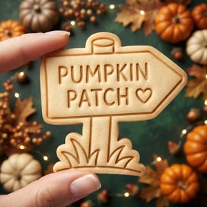 May include: A light beige, arrow-shaped cookie with embossed text reading PUMPKIN PATCH and a small heart symbol. The cookie features a post design with stylized grass at the base, showcasing a smooth, baked surface with crisp, defined edges.