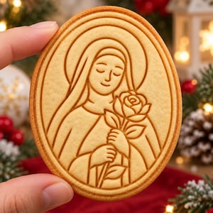 May include: An oval-shaped light brown cookie featuring an embossed line drawing of a figure with a halo, wearing a head covering and holding a single rose. The design is centered within a double-lined oval border on the surface of the baked treat.