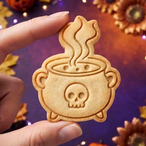 May include: A light beige cookie shaped like a cauldron with a skull emblem on the front and steam rising from the top. The cookie features embossed details including a round pot, two side handles, three small feet, and a skull with eye sockets.