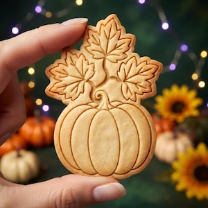 May include: A light tan, baked biscuit shaped like a pumpkin with three maple leaves extending from the top. The biscuit features embossed lines detailing the pumpkin's segments and the veins of the leaves, with a smooth, flat surface texture.