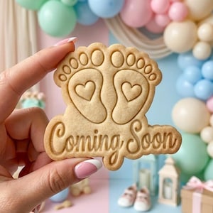 May include: A light brown cookie in the shape of baby footprints with hearts and the words "Coming Soon" in a decorative font. The cookie is held in front of a colorful balloon backdrop.
