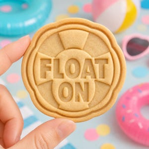 May include: A round, light brown cookie with a scalloped edge, featuring a raised design of a life preserver and the embossed text "FLOAT ON" in the center. The cookie has a smooth, baked texture and a distinct, molded relief pattern.