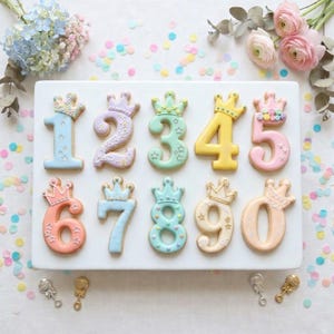 May include: A collection of ten number-shaped cookies, each adorned with a crown. The cookies are decorated with pastel colors including blue, pink, yellow, and green. The cookies are arranged on a white rectangular platter, with confetti and flowers in the background.