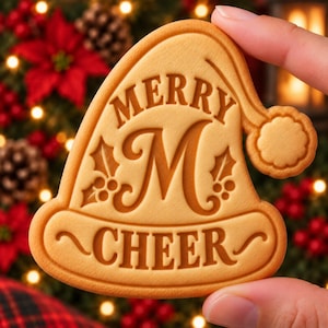 May include: A light tan biscuit shaped like a Santa hat with the embossed text MERRY CHEER and a large letter M in the centre. The design includes holly leaf and berry accents on both sides of the letter M and a textured pom-pom on the hat tip.