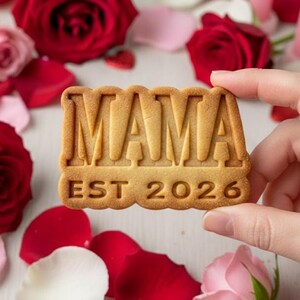 May include: A rectangular cookie with the word "MAMA" embossed on top and "EST 2026" below. The cookie is held by a hand, surrounded by red and pink rose petals and roses.