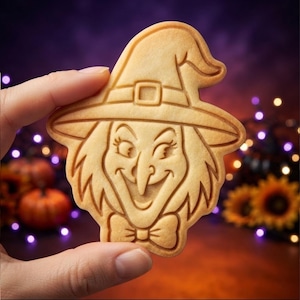 May include: A light tan, flat, baked cookie shaped like a witch's head wearing a pointed hat with a buckle and a bow tie at the neck. The cookie features embossed facial details including eyes, a nose, and a wide, smiling mouth.
