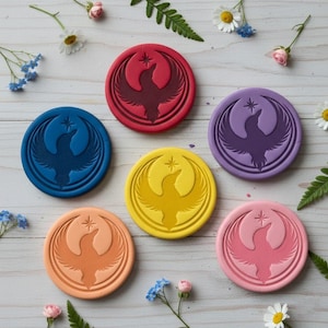 May include: A collection of six colorful, circular coasters. Each coaster features a stylized bird design with a star, in shades of blue, red, purple, yellow, orange, and pink. The coasters are arranged on a light-colored surface, decorated with flowers and greenery.