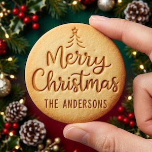 May include: A round, light brown cookie featuring an embossed design of a small Christmas tree above the cursive text Merry Christmas and the block letter text THE ANDERSONS, centered on the surface of the baked treat.