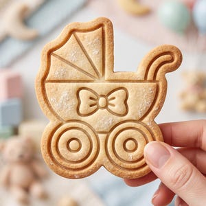 May include: A light brown, baked cookie shaped like a baby carriage. The cookie features embossed details including a canopy with lines, a central bow tie design, and two circular wheels. The surface has a slightly textured, golden-brown finish.