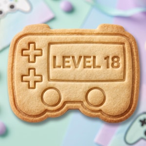 May include: A light brown, rectangular-shaped cookie shaped like a video game controller. The surface features an embossed design of a screen with the text LEVEL 18, a directional pad with two plus signs, and two circular buttons at the bottom.