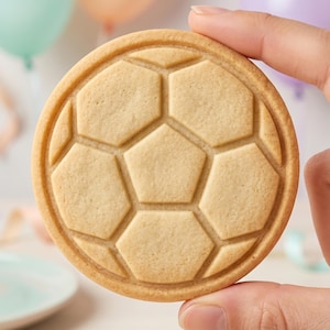 May include: A round, light tan sugar cookie featuring an embossed geometric pattern of hexagons and a central pentagon, mimicking the classic design of a soccer ball. The cookie has a smooth, baked texture with defined edges and a uniform color.