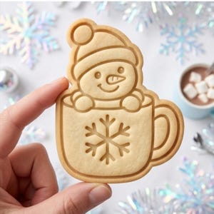 May include: A light tan cookie cutter shaped like a snowman wearing a hat, sitting inside a mug decorated with a snowflake design. The cookie cutter features embossed details including the snowman's face, hat, and the snowflake on the mug.