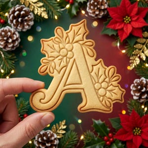 May include: A light tan, letter-shaped cookie featuring an embossed design of holly leaves and berries. The cookie is shaped like the capital letter A, with a smooth surface and defined edges, showcasing a detailed botanical pattern.
