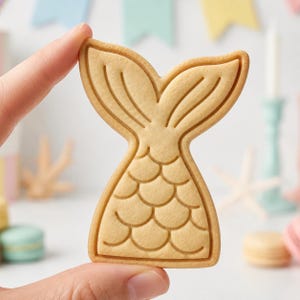 May include: A light tan, baked sugar cookie shaped like a mermaid tail. The cookie features embossed details including a scalloped scale pattern on the lower section and defined lines on the two-pronged tail fin at the top, with a smooth texture.