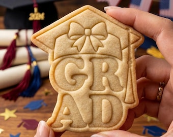 May include: A graduation-themed cookie in the shape of a mortarboard cap with the word "GRAD" in the center. The cookie is held in a person's hand. Graduation caps, diplomas, and American flags are in the background.