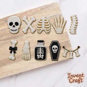 May include: A set of nine skeleton-themed cookies featuring a skull, crossbones, rib cage, hand, spine, bone with a black bow, foot, potion bottle with a skeleton graphic, and a coffin with a skull, plus a small sign reading Invent Craft.