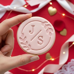 Peut inclure: Un biscuit rond rose clair avec les initiales "J/S" à l'intérieur d'une couronne de laurier. Le biscuit est tenu sur un fond rouge avec des coeurs et des rubans décoratifs. Le biscuit a un bord brun doré.