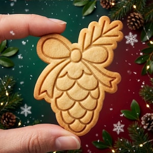 May include: A light brown, baked cookie shaped like a pinecone with a decorative bow at the top and a sprig of needles extending from the side. The cookie features embossed lines that define the scales of the pinecone and the texture of the bow.