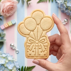 May include: A light beige sugar cookie shaped like a gift box with a large bow and three balloons rising from the top. The cookie features embossed details including polka dots on the box and defined outlines for the balloons and ribbon.