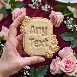 May include: A light-colored cookie with the words "Any Text" embossed on it. The cookie is rectangular with rounded corners and floral designs. Pink roses and greenery surround the cookie on a burgundy surface.