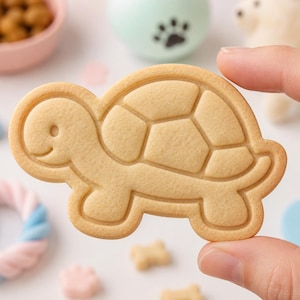 May include: A light tan, turtle-shaped cookie with embossed details outlining the head, shell, and legs. The cookie has a smooth, matte surface texture and a distinct, raised border that defines the shape of the turtle, including a small eye and mouth.