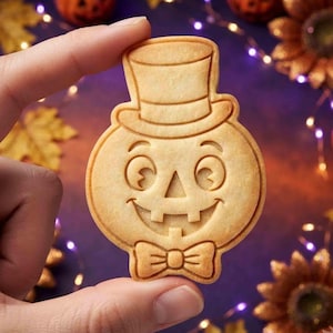 May include: A light tan, round-shaped cookie featuring an embossed design of a jack-o'-lantern wearing a top hat and a bow tie. The cookie has a smooth, baked texture with clearly defined lines depicting the face, hat, and bow tie details.
