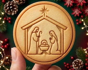 May include: A round, light brown cookie featuring an embossed nativity scene. The design depicts two figures standing and kneeling beside a central manger, all sheltered under a stable roof with a prominent star positioned at the very top peak.