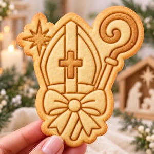 May include: A light tan, baked sugar cookie shaped like a mitre hat with a cross, a star, and a bishop's crozier staff. The cookie features embossed lines creating a detailed design with a bow at the base of the mitre and a scalloped edge.