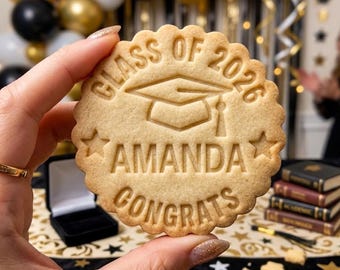 May include: A round, light-brown cookie with a scalloped edge. The cookie is stamped with the words "CLASS OF 2026", a graduation cap, the name "AMANDA", and the word "CONGRATS". The cookie is held in front of a blurred background.