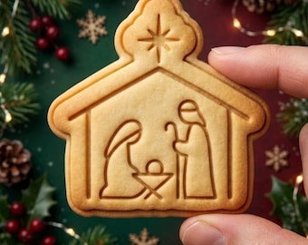 May include: A light tan, house-shaped sugar cookie featuring an embossed nativity scene. The design depicts a central star above a stable containing a figure kneeling beside a manger and a standing figure holding a staff, all rendered in clean, simple lines.
