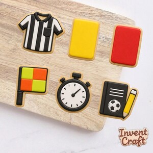 May include: A set of six decorated cookies shaped like soccer referee gear, including a black and white striped shirt, a yellow card, a red card, a checkered flag, a stopwatch, and a black notebook with a soccer ball and a yellow pencil.