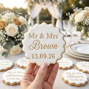 May include: A white, scalloped-edged cookie with gold lettering that reads "Mr & Mrs Brown 13.09.26" is held in front of a table setting. Other cookies with the same design are scattered on the table, along with floral arrangements and glassware.