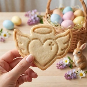 May include: A baked angel-shaped cookie with a heart and cross design. The cookie is held in front of a basket of pastel-colored Easter eggs, a ceramic bunny, and sprigs of purple flowers. The cookie has a halo and wings.