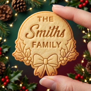 May include: A round, light brown cookie featuring an embossed design of a decorative bow and holly leaves at the bottom. The text THE Smiths FAMILY is centered on the cookie in a combination of serif and script fonts, creating a festive look.